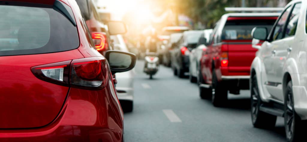 A red car in heavy traffic with a blurred motorcycle passing between lanes in the background.