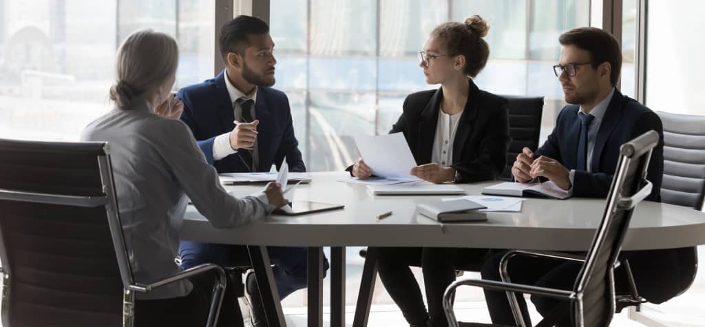 A group of four professionals in business attire engaged in a discussion around a modern table in a well-lit office.