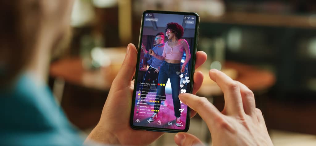 A person holds a smartphone, viewing a TikTok video of a dancer in vibrant attire, surrounded by colorful lights and engaging content.