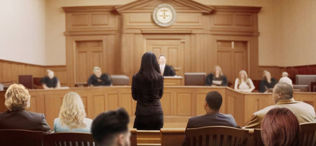 A courtroom scene featuring a woman speaking, with judges and jurors attentively listening from their seats. The wooden interior is prominent.