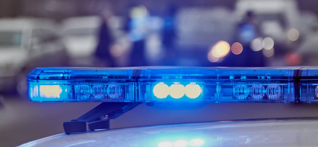 A close-up of a police car's blue emergency light, with blurred police activity and vehicles in the background.