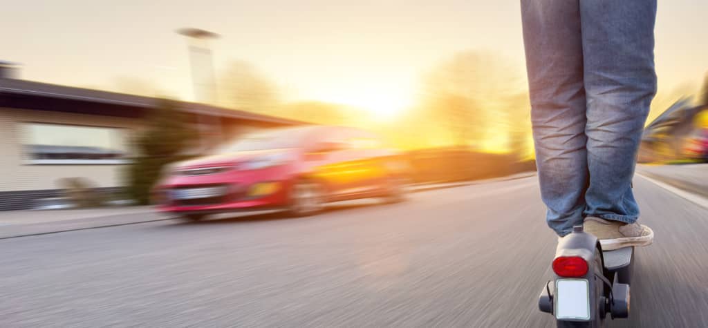 A person rides a scooter down a road as a car speeds past, with a sunset glowing in the background.
