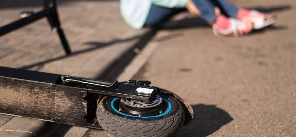 A fallen electric scooter lies on the ground near a person sitting on the pavement, visibly distressed after an accident.