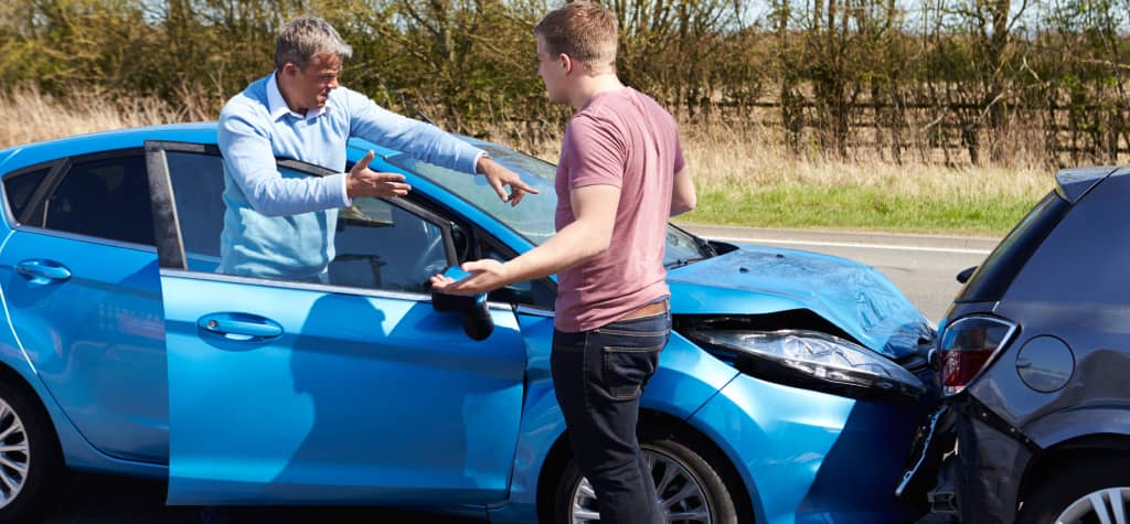 Two men are discussing a minor car accident, with one man gesturing towards the damage on the blue car parked on the roadside.
