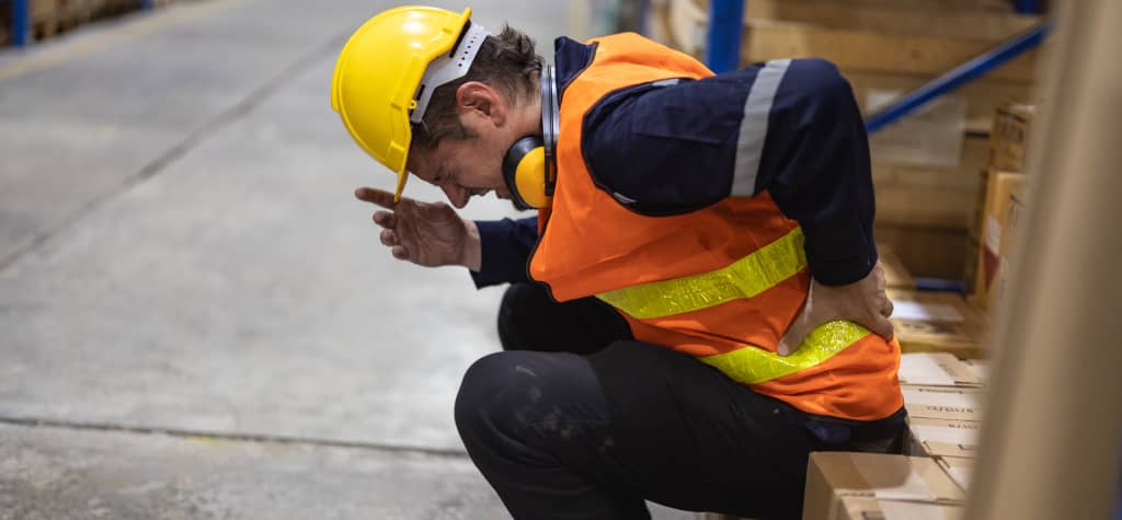 A warehouse worker in a yellow hard hat and orange vest is crouched, holding his lower back in discomfort amidst stacked boxes.