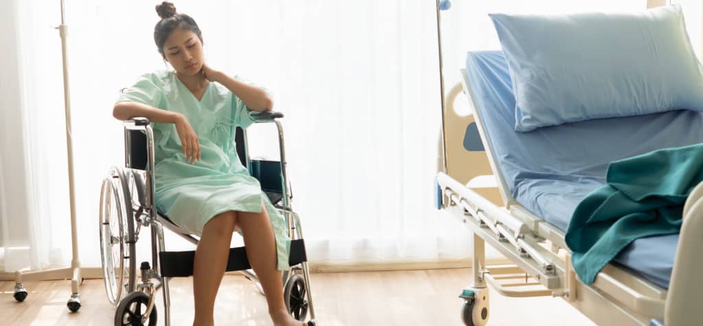 A person in a hospital gown sits in a wheelchair beside an unoccupied hospital bed, with natural light filtering through the window.