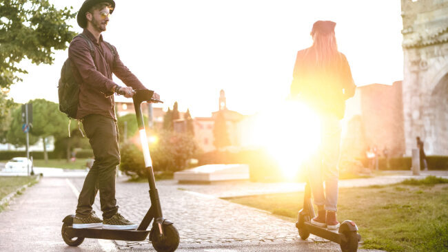 Man and woman riding e-scooters along a brick sidewalk on a college campus near sunset.