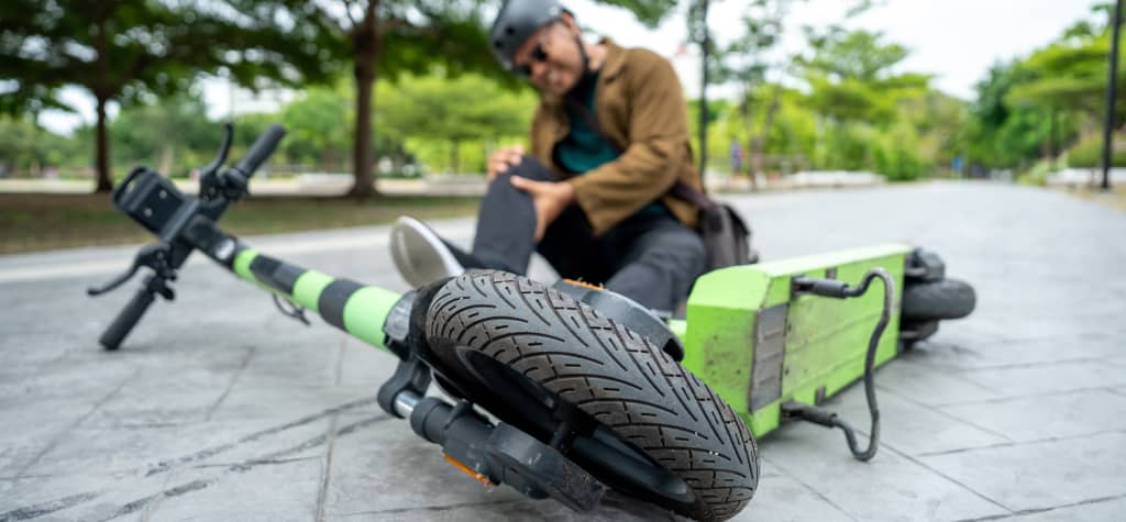 A person in a helmet is seated on the ground next to a fallen green and black electric scooter, holding their knee on a park pathway.