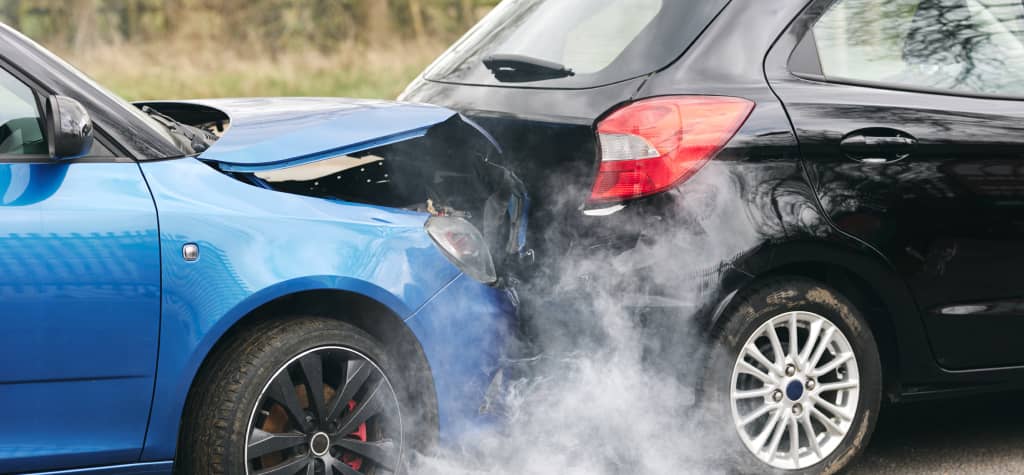 A blue car with a crumpled hood collided with a black car, emitting smoke from the impact site.