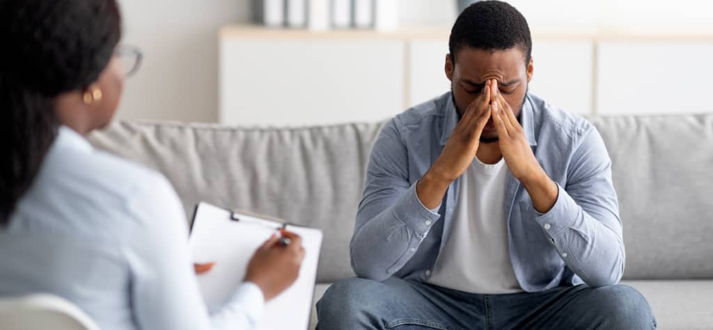 A therapist takes notes while a client sits on a couch, looking contemplative and engaged in a serious conversation.