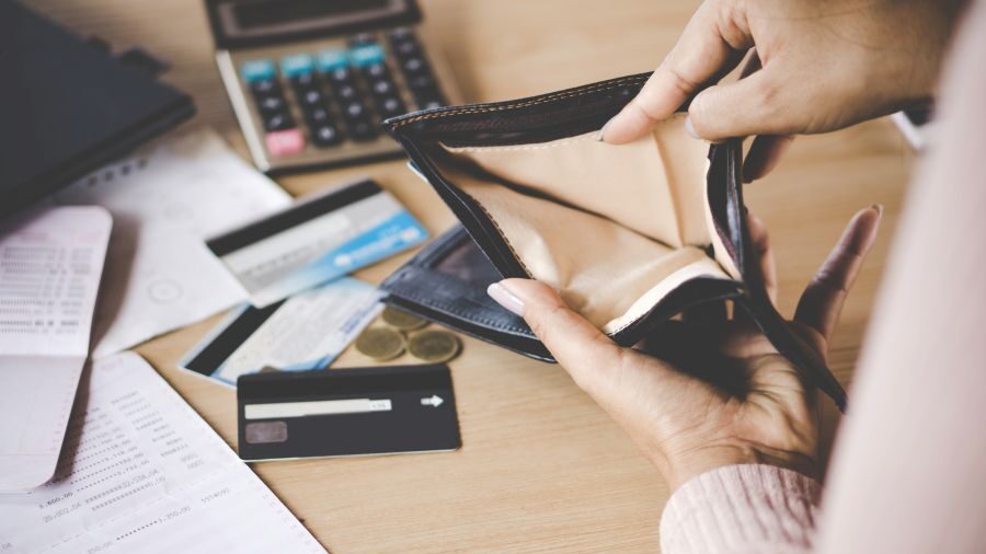 Two hands holding an empty wallet above a desk with papers, credit cards, coins, and a calculator scattered on a desk.