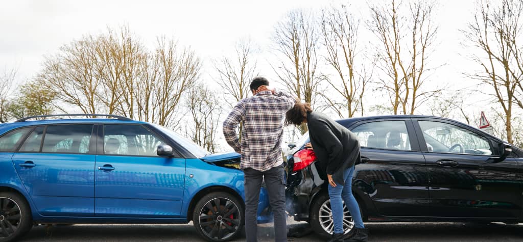 Two people inspect damage after a minor car accident involving a blue and a black vehicle, with trees visible in the background.