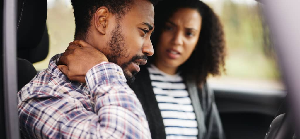 A man in a plaid shirt sits in a car, rubbing his neck, while a woman beside him sports curly hair and a striped shirt.