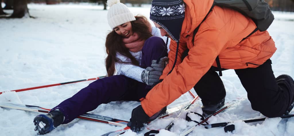 A person in an orange jacket assists someone sitting in the snow, surrounded by skiing equipment, in a snowy outdoor setting.