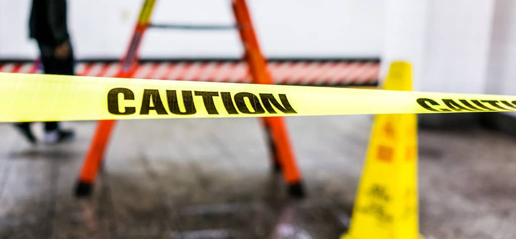Caution tape surrounds a ladder and a warning cone in a work area, indicating maintenance or potential hazards. A person walks nearby.