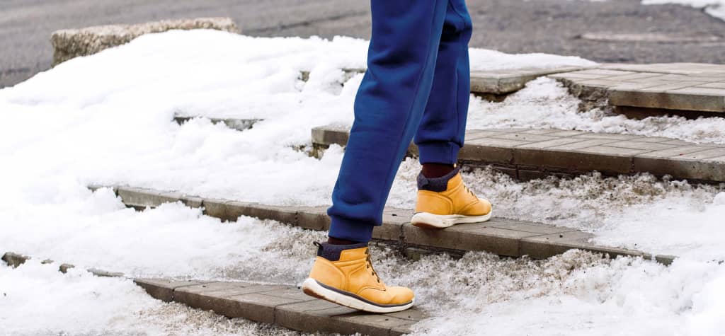 A person in blue sweatpants and yellow sneakers walks down steps covered in snow and slush.